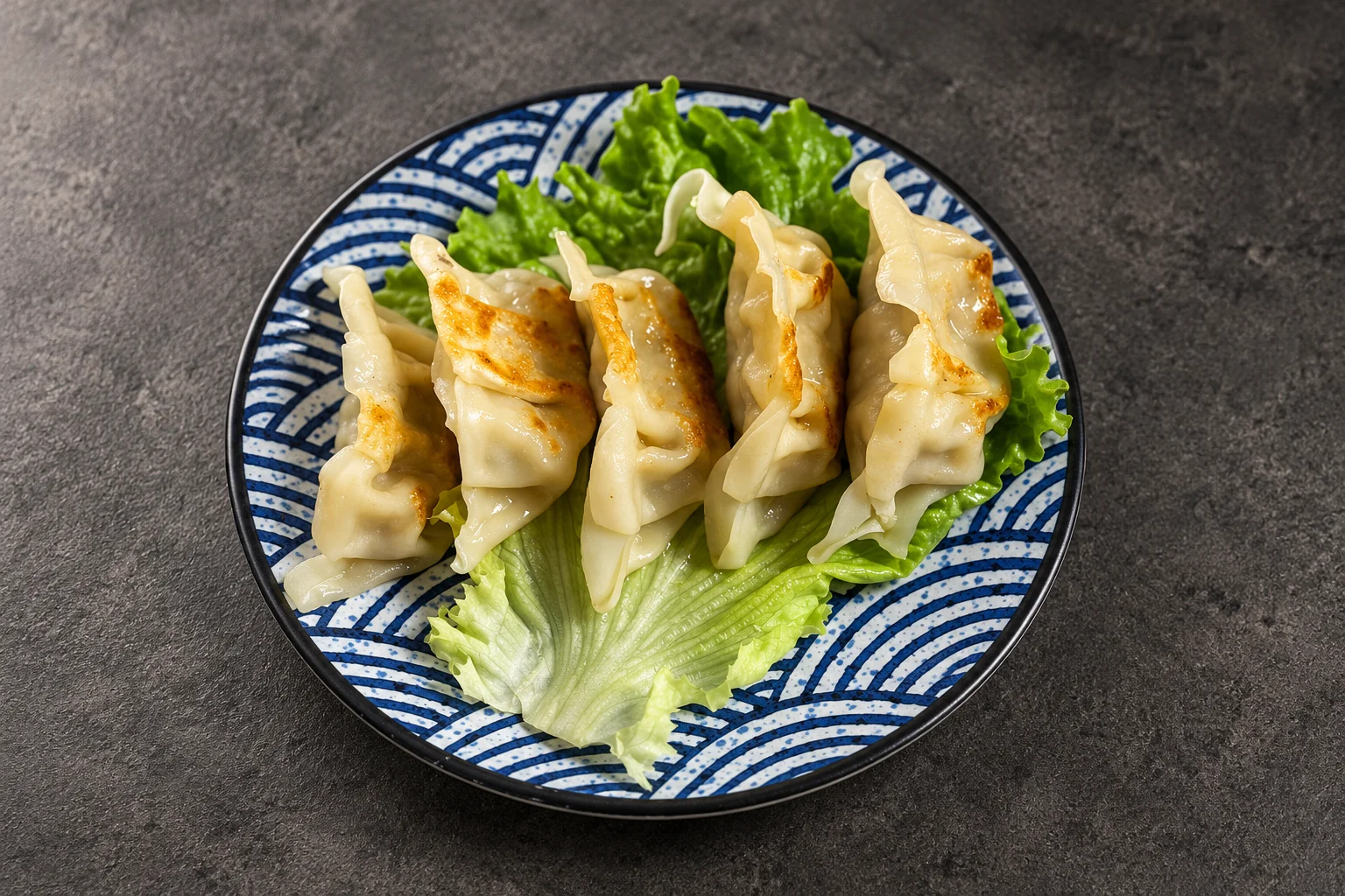 Gyoza sur feuille de salade, assiette à motif de vagues bleues, fond de béton sombre.